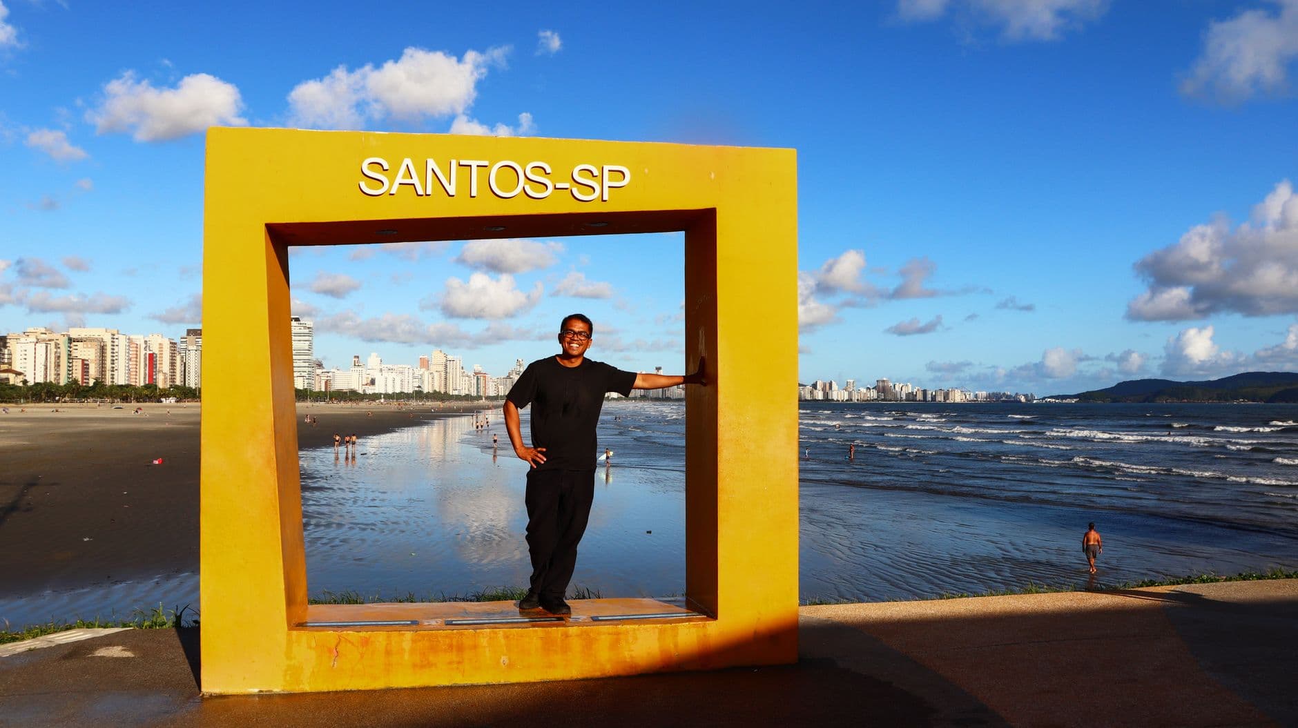 Person posing at Santos-SP landmark with city skyline and beach view.