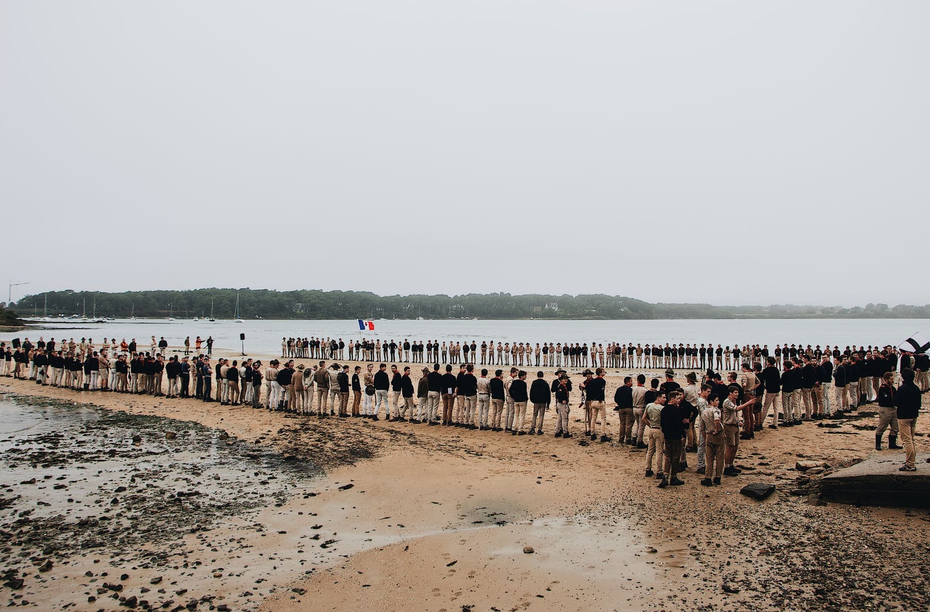 A large group of scouts gathered on a seashore in daylight, forming a circle with flags.