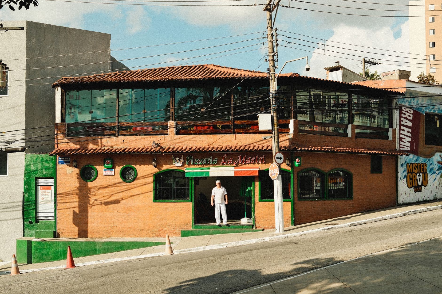 A sunny corner view of a quaint pizzeria, featuring a lone man standing in the entrance, under a clear blue sky.