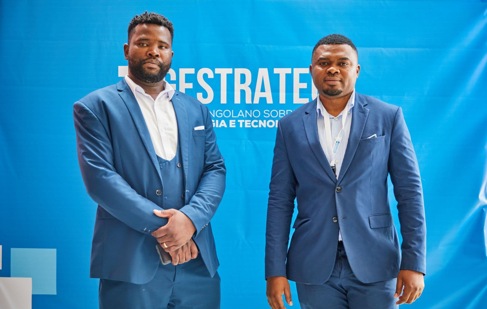 Two men in blue suits pose at a professional conference against a branded backdrop.